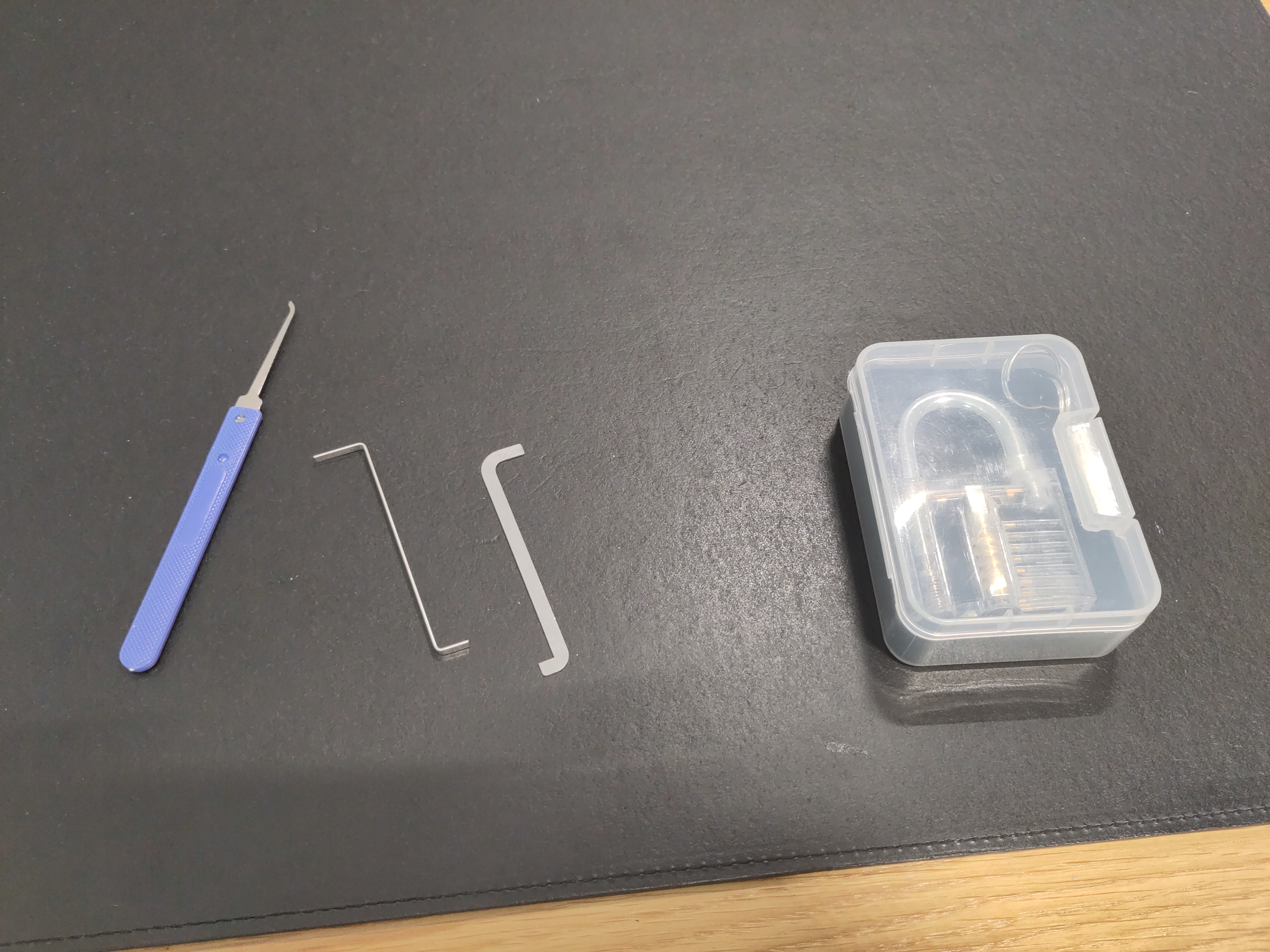 A lockpick and two tensioners on a table, along with a plastic box with a see-through training lock inside. 