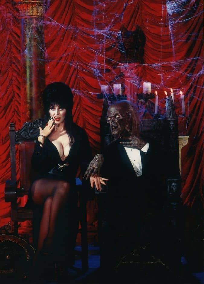 Elvira sitting beside the Cryptkeeper with her hand to her cheek, and his hand on her left hand on an armrest.