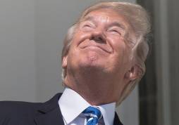 The image shows former U.S. President Donald Trump looking up at the sky, squinting with a slight smile. He is wearing a dark suit with a blue-striped tie, and his face is tilted upward, giving him a look of curiosity or amusement. The background suggests an outdoor setting, likely near a building or structure.