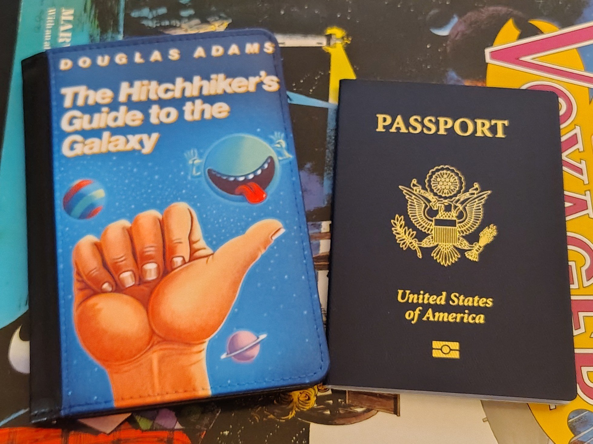 Photo of a US passport and a passport cover custom-printed with the cover art from the original US edition of the novel "The Hitchhiker's Guide to the Galaxy." Said art features a hand giving the universal thumb gesture for hitching a ride while a green planet makes a silly face against a backdrop of space. (Douglas Adams and many fans dislike this version of the cover, but it was on the first copy of the story I had as a kid and I'll always have great fondness for it.)