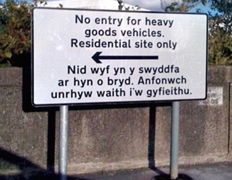 Sign on a road. "No entry for heavy goods vehicles. Residential site only. (Heavy arrow pointing left) Nid wyf yn y swyddfa ar hyn o bryd. Anfonwch unrhyw waith i'w gyfieithu."