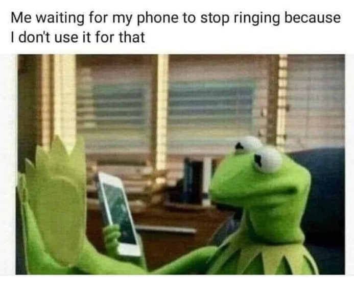 A photograph of the hand puppet "Kermit the frog" inside an office with a smart phone. There is text above the photograph. Kermit is implied to be sitting on a computer chair, casually resting his legs up as if leaning them on a desk. Kermit is holding the phone and looking it's screen. The text above reads, "me waiting for my phone to stop ringing as I don't use it for that".