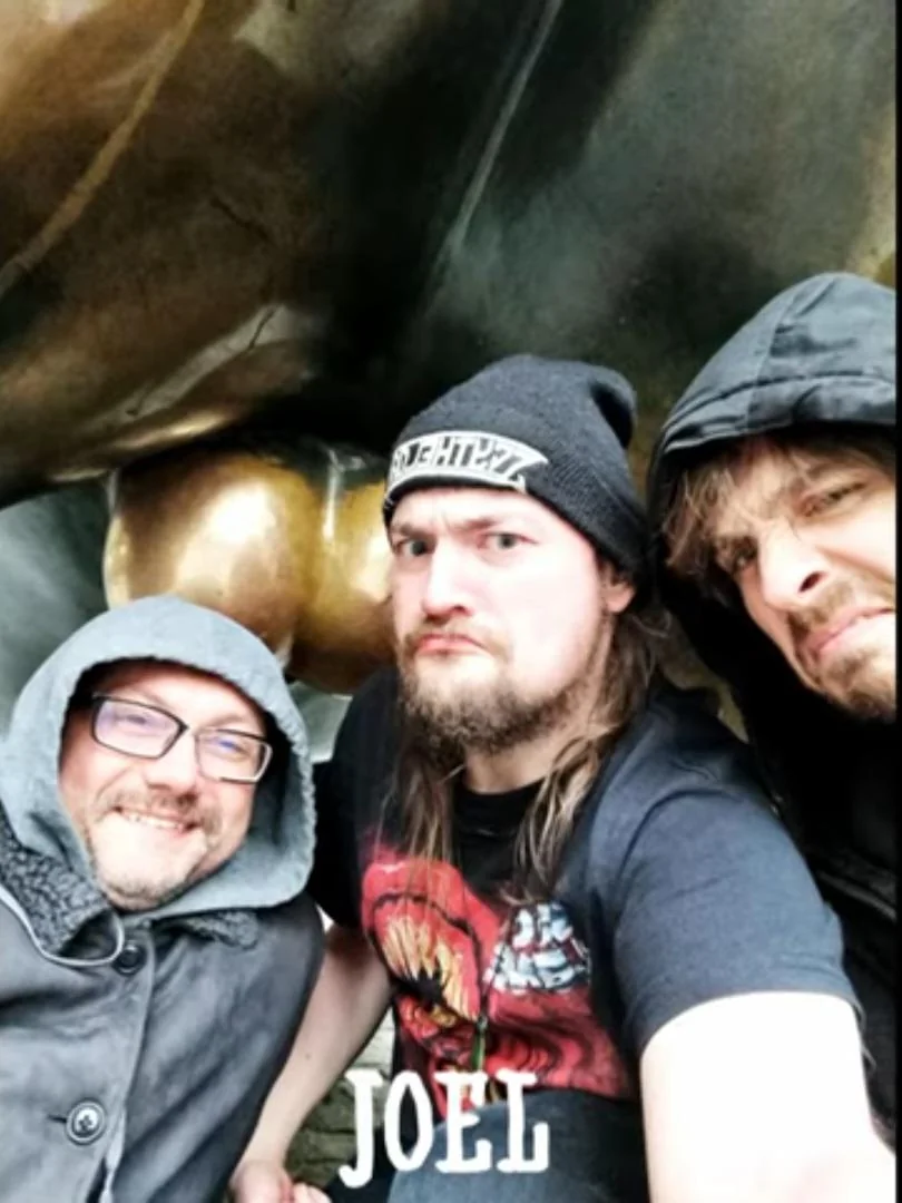 A picture of Vinny Vinesauce, Vargskelethor Joel, and Jabroni Mike in front of a large, well-polished pair of testicles on a statue.